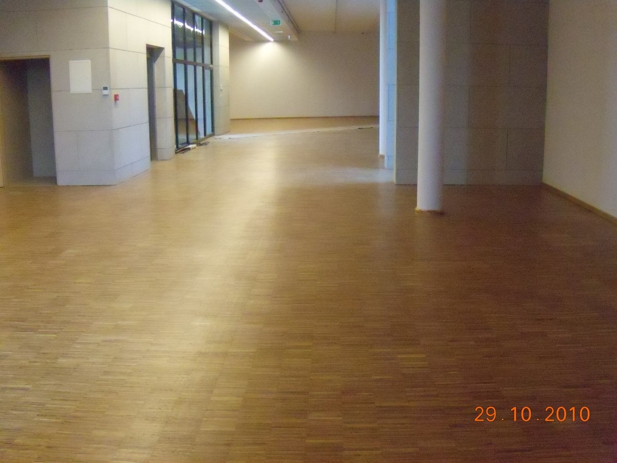 Wnętrze komercyjne z parkietem ułożonym w jodełkę, jasne ściany z kolumnami i widoczne drzwi oraz okna w oddali.