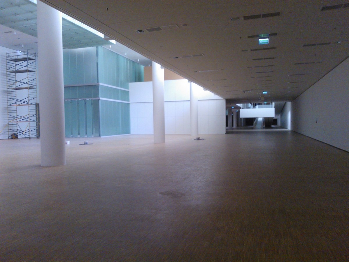 Przestronne wnętrze w trakcie wykończenia, z białymi kolumnami, szklanymi ścianami i rozstawionym rusztowaniem w rogu, ukazujące postęp prac.