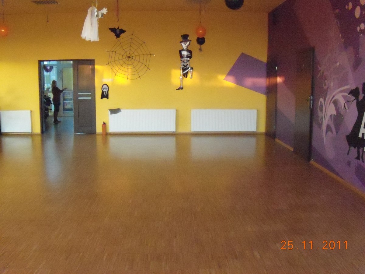 Pusta sala z żółtymi ścianami udekorowana na Halloween, widoczne dekoracje: duch, pająk, szkielet w cylindrze, pod ścianami zamontowane białe grzejniki, podłoga parkietowa