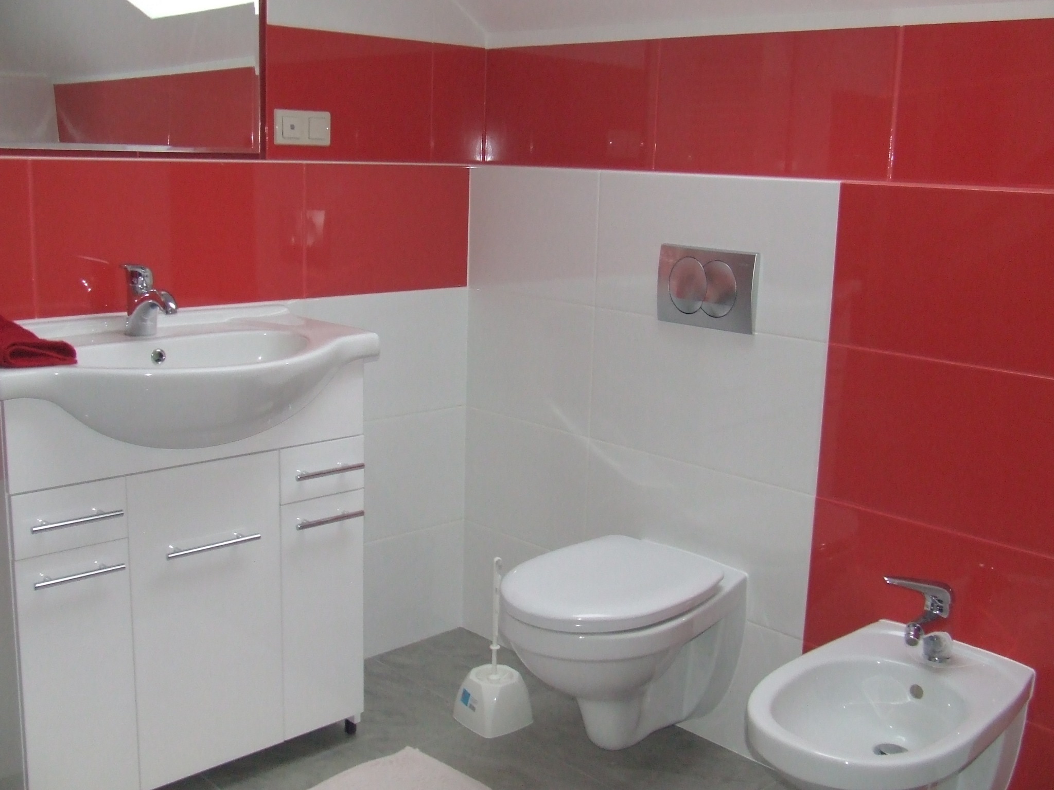 Biało-czerwona łazienka z umywalką na szafce, bidetem i toaletą podwieszaną, wykończona błyszczącymi płytkami.