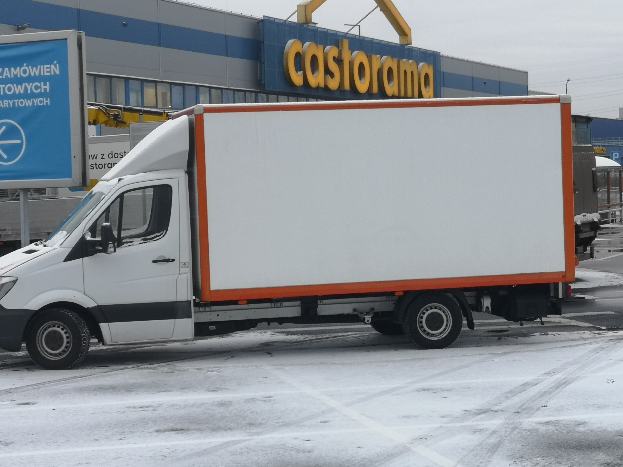 Biały samochód dostawczy z pomarańczowymi obramowaniami zaparkowany na ośnieżonym parkingu przed budynkiem Castoramy.