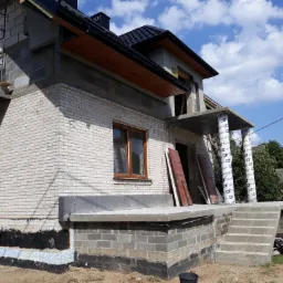 Surowy stan domu jednorodzinnego w trakcie rozbudowy: widoczne białe cegły, niezadaszony taras z kolumnami owiniętymi folią, fragment ocieplenia fundamentów oraz niebieskie niebo w tle.