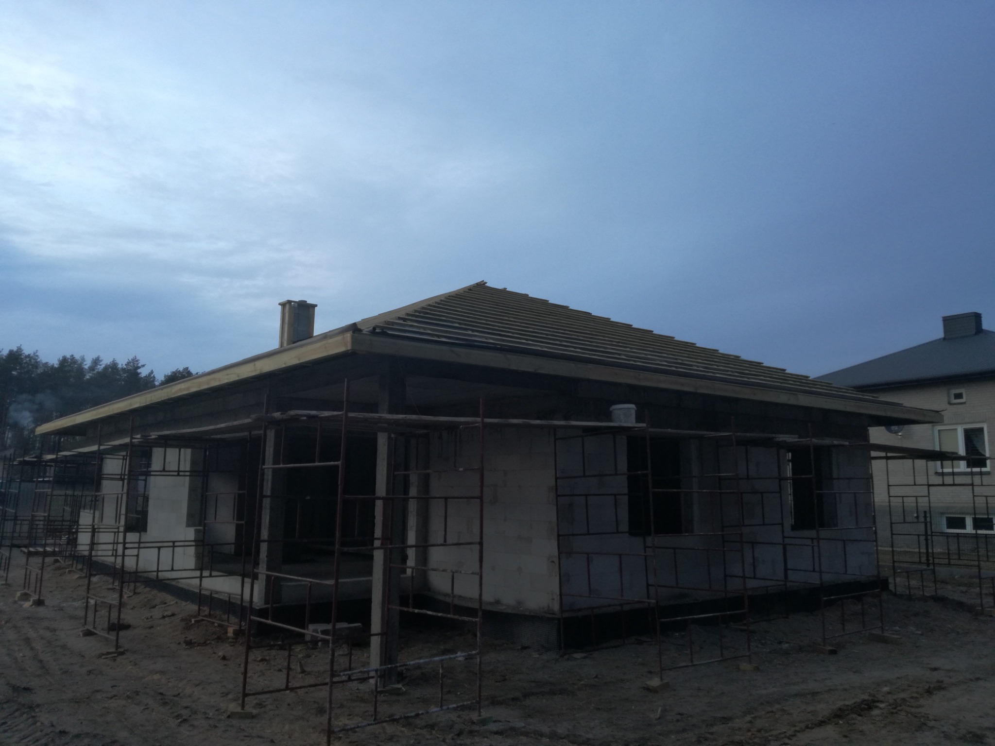 Budowa domu jednorodzinnego w stanie surowym otwartym, widoczne szare bloczki, drewniana konstrukcja dachu i rusztowania, pochmurne niebo w tle.