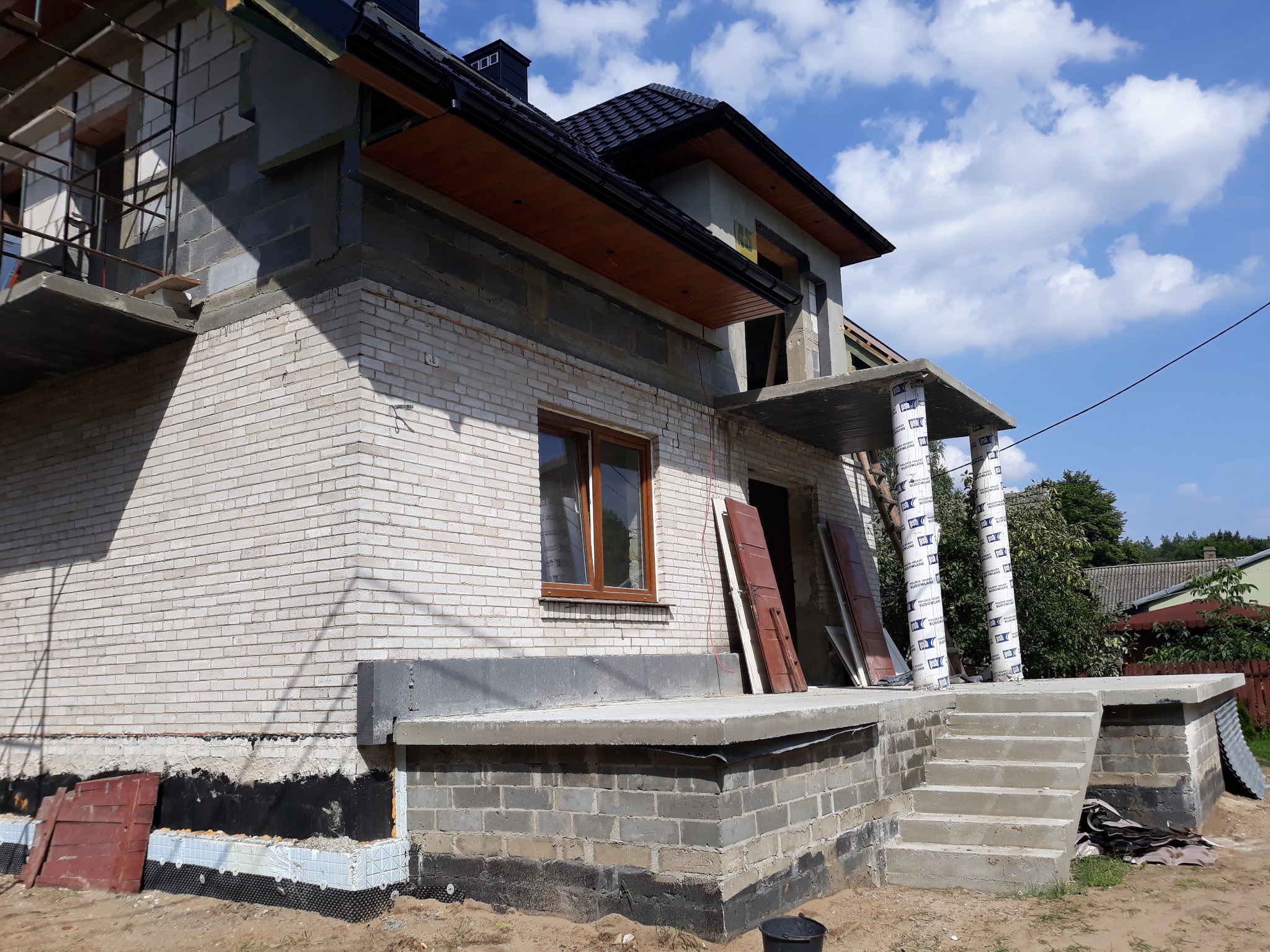 Surowy stan domu jednorodzinnego w trakcie rozbudowy: widoczne białe cegły, niezadaszony taras z kolumnami owiniętymi folią, fragment ocieplenia fundamentów oraz niebieskie niebo w tle.