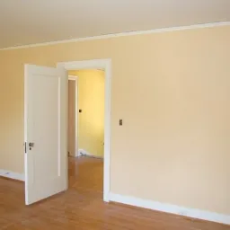 Jasny pokój z pomalowanymi na kremowo ścianami, drewnianą podłogą i białymi drzwiami, widok na sąsiednie pomieszczenie z żółtą ścianą w tle.