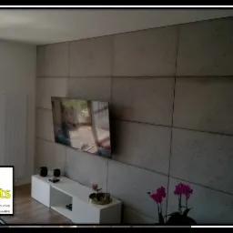 Ściana wykończona betonowymi panelami w salonie w Kielcach, z wiszącym telewizorem i białym regałem pod spodem, ozdobiona kwiatami.