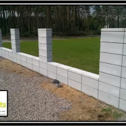 Wznoszony mur z szarej kostki betonowej, widoczny na tle zielonego trawnika i lasu, z logo firmy budowlanej w lewym dolnym rogu.