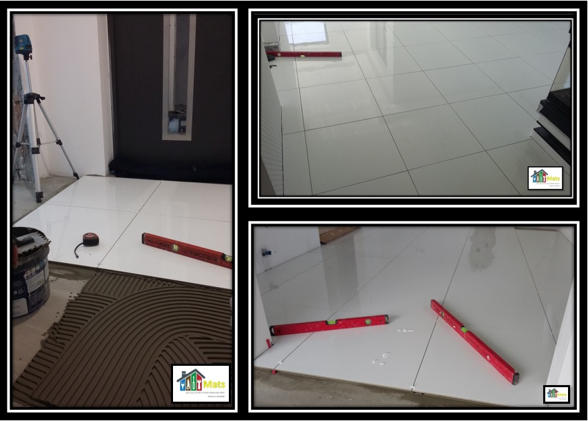 Układanie białych, błyszczących płytek podłogowych w przedpokoju, widoczny laser krzyżowy na statywie, poziomica, puszka z klejem i grzebień do rozprowadzania kleju na podłodze.