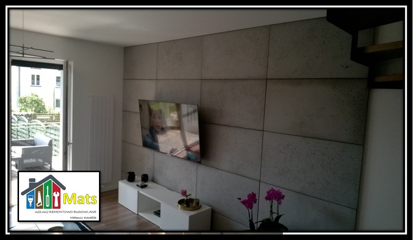 Ściana wykończona betonowymi panelami w salonie w Kielcach, z wiszącym telewizorem i białym regałem pod spodem, ozdobiona kwiatami.