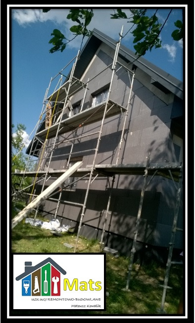 Dom jednorodzinny w trakcie ocieplania, widoczne rusztowania i grafitowe płyty termoizolacyjne na ścianach, w dolnym rogu logo firmy remontowo-budowlanej.