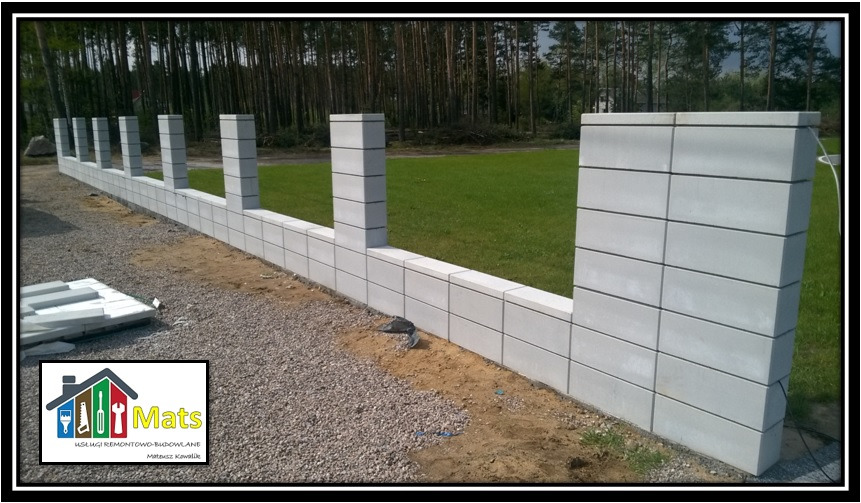 Wznoszony mur z szarej kostki betonowej, widoczny na tle zielonego trawnika i lasu, z logo firmy budowlanej w lewym dolnym rogu.