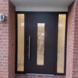 Ciemna, nowoczesna brama wejściowa z długim, pionowym uchwytem i szybami z ornamentowego szkła, wkomponowana w czerwoną ceglaną fasadę z numerem domu na boku.