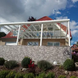 Zadaszenie tarasu z białym stelażem i przeszklonym dachem, widok z dołu na tle domu z czerwonym dachem i błękitnego nieba z chmurami.