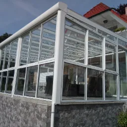 Zewnętrzny widok przeszklonej werandy z białymi ramami okiennymi, wewnątrz widoczne rattanowe meble ogrodowe, podmurówka wykończona kamieniem elewacyjnym.