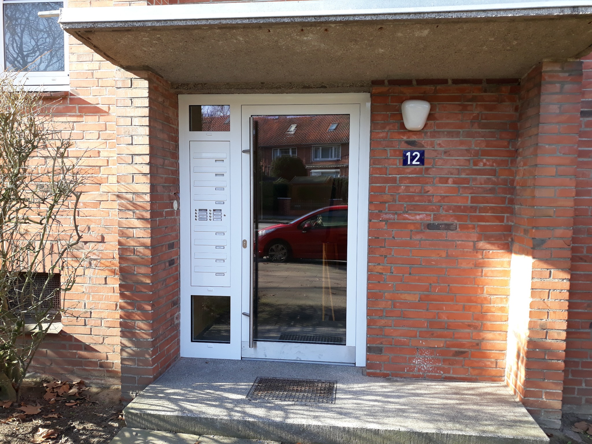 Wejście do budynku z białymi drzwiami z wbudowanymi skrzynkami pocztowymi, widoczny numer 12 na ceglanej ścianie i odbicie czerwonego samochodu w szybie.