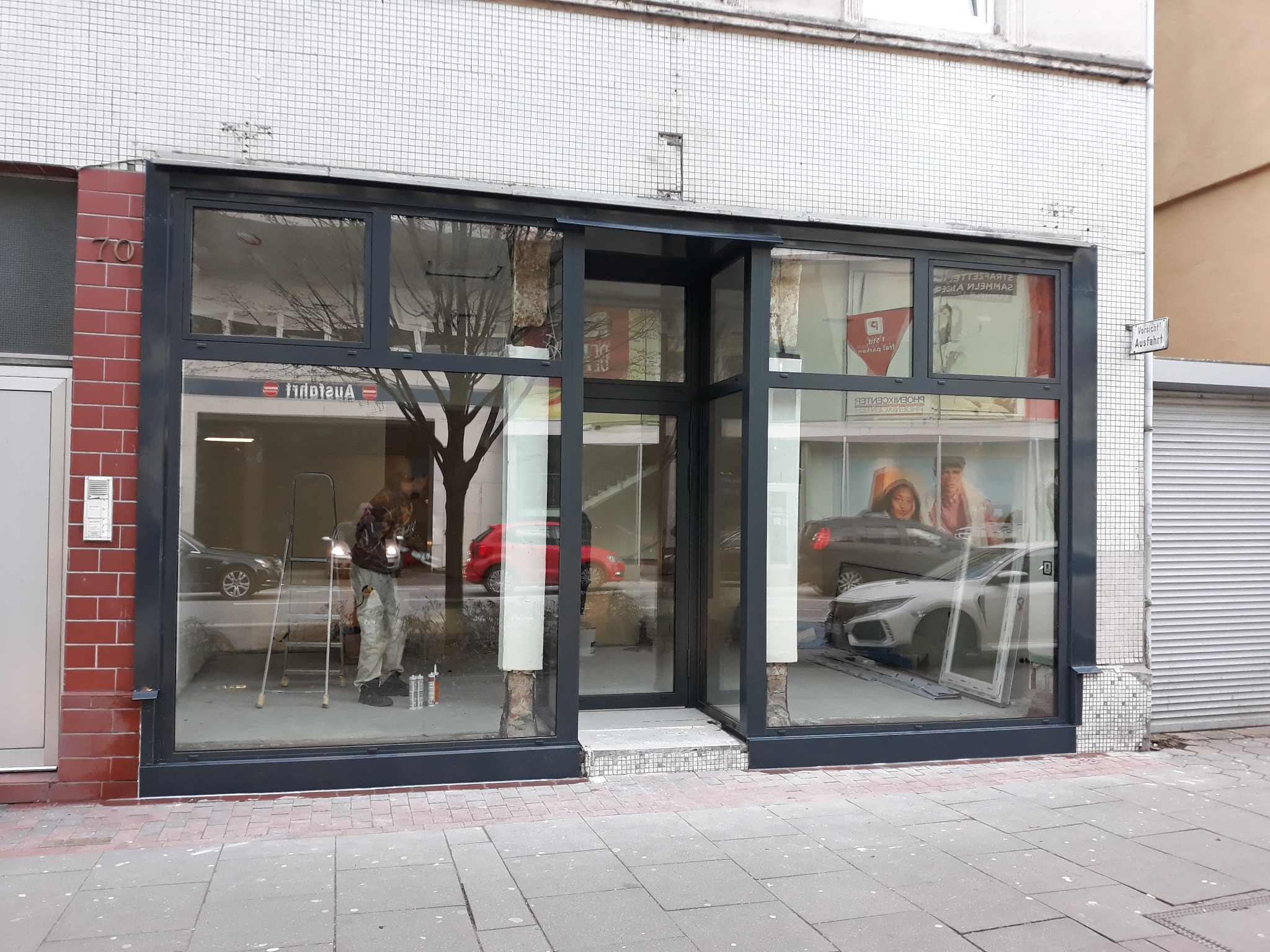 Czarny szkielet witryny sklepowej z widocznym pracownikiem na drabinie, przygotowującym się do montażu szyb, z ulicą i samochodami odbijającymi się w szybach.