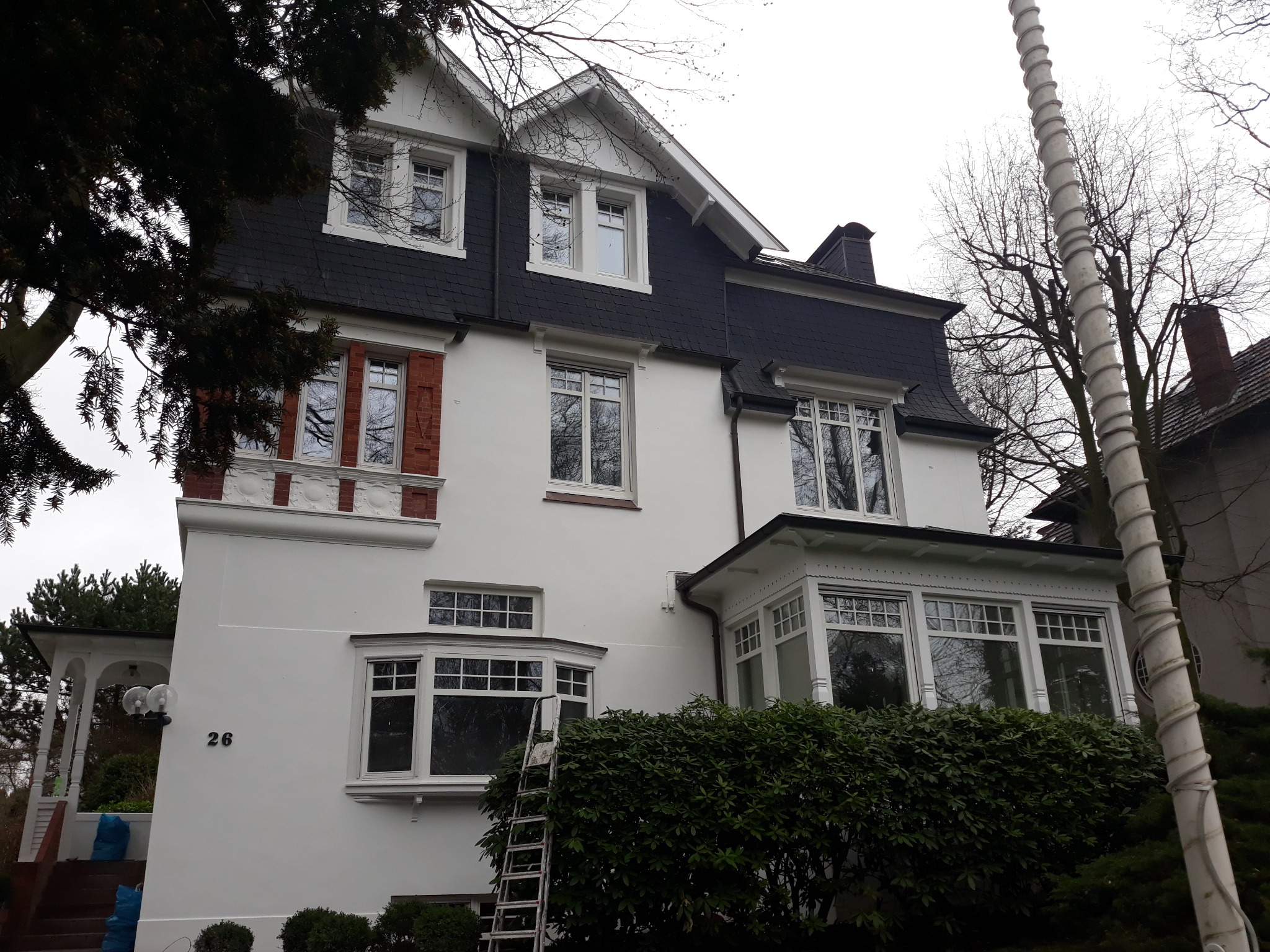 Elewacja dwupiętrowego domu z białymi ramami okiennymi, czarnym dachem z dachówki, numerem 26 na ścianie i metalową drabiną opartą o ścianę, częściowo zasłoniętą przez krzew.