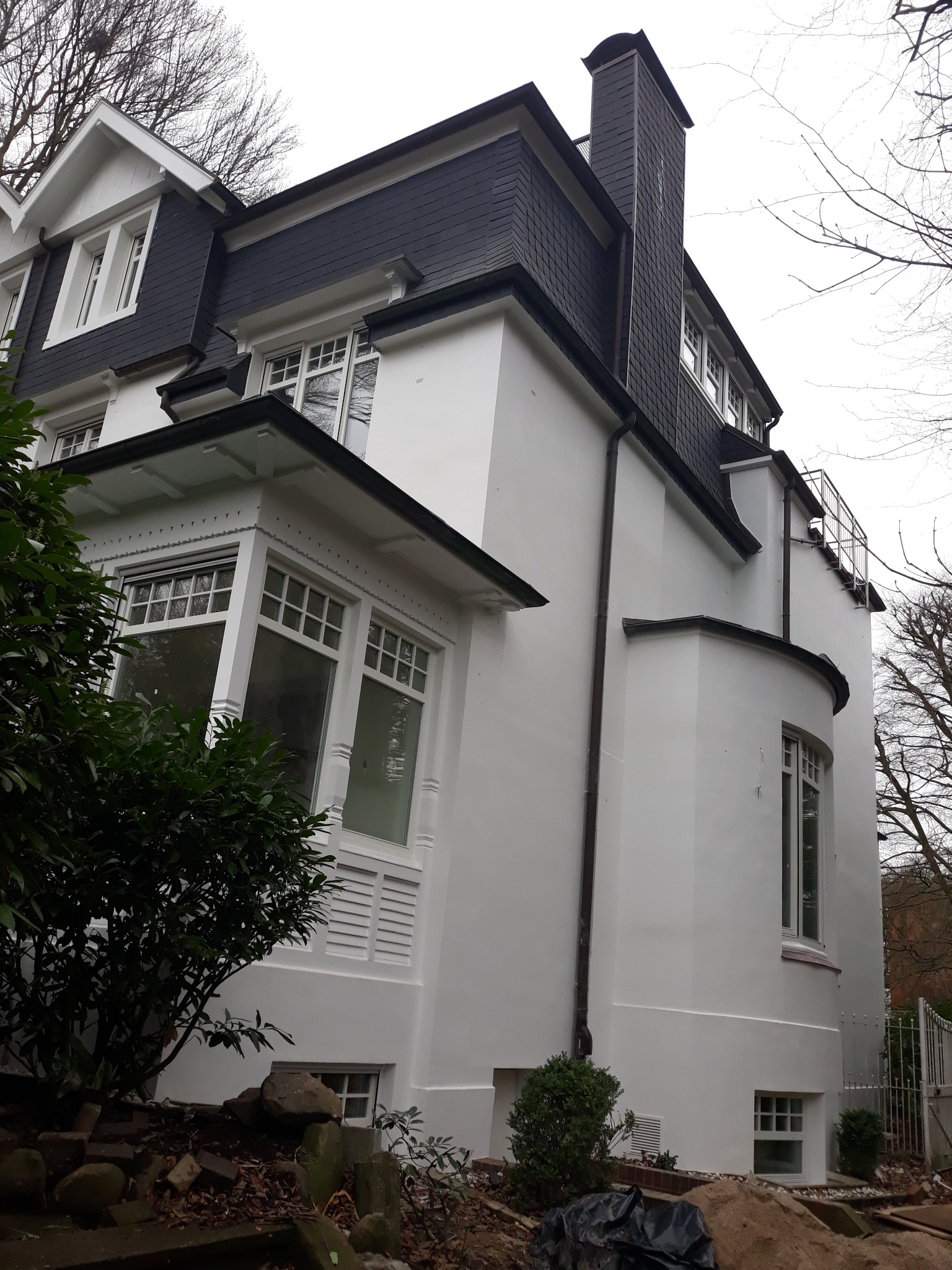 Elewacja odnowionego, białego domu z ciemnym dachem z dachówki, widok z dołu, z fragmentem ogrodu z przodu.