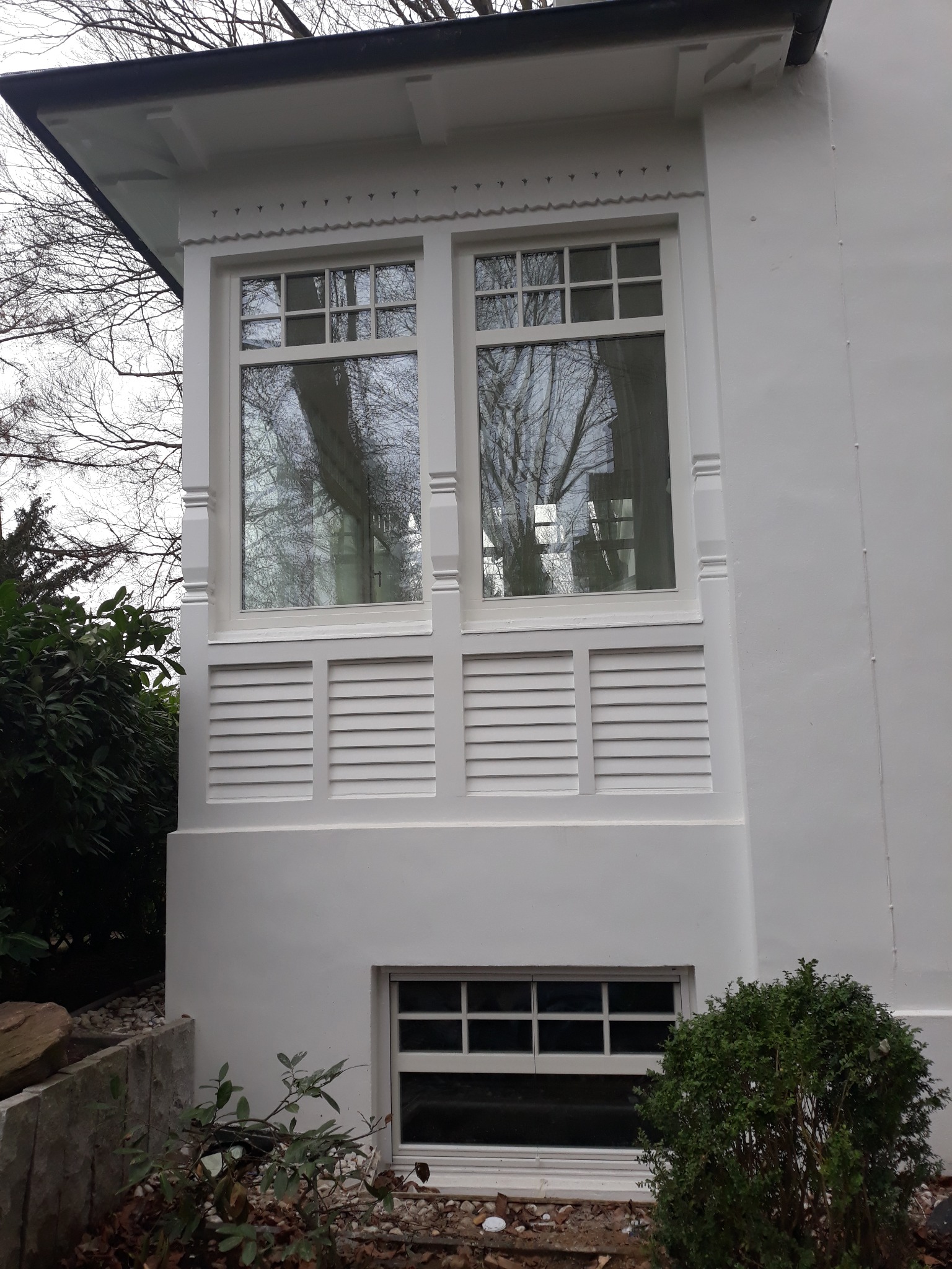 Elewacja budynku z białymi oknami z szprosami i ozdobnymi elementami architektonicznymi, odbicie drzew w szybach, widok z zewnątrz.