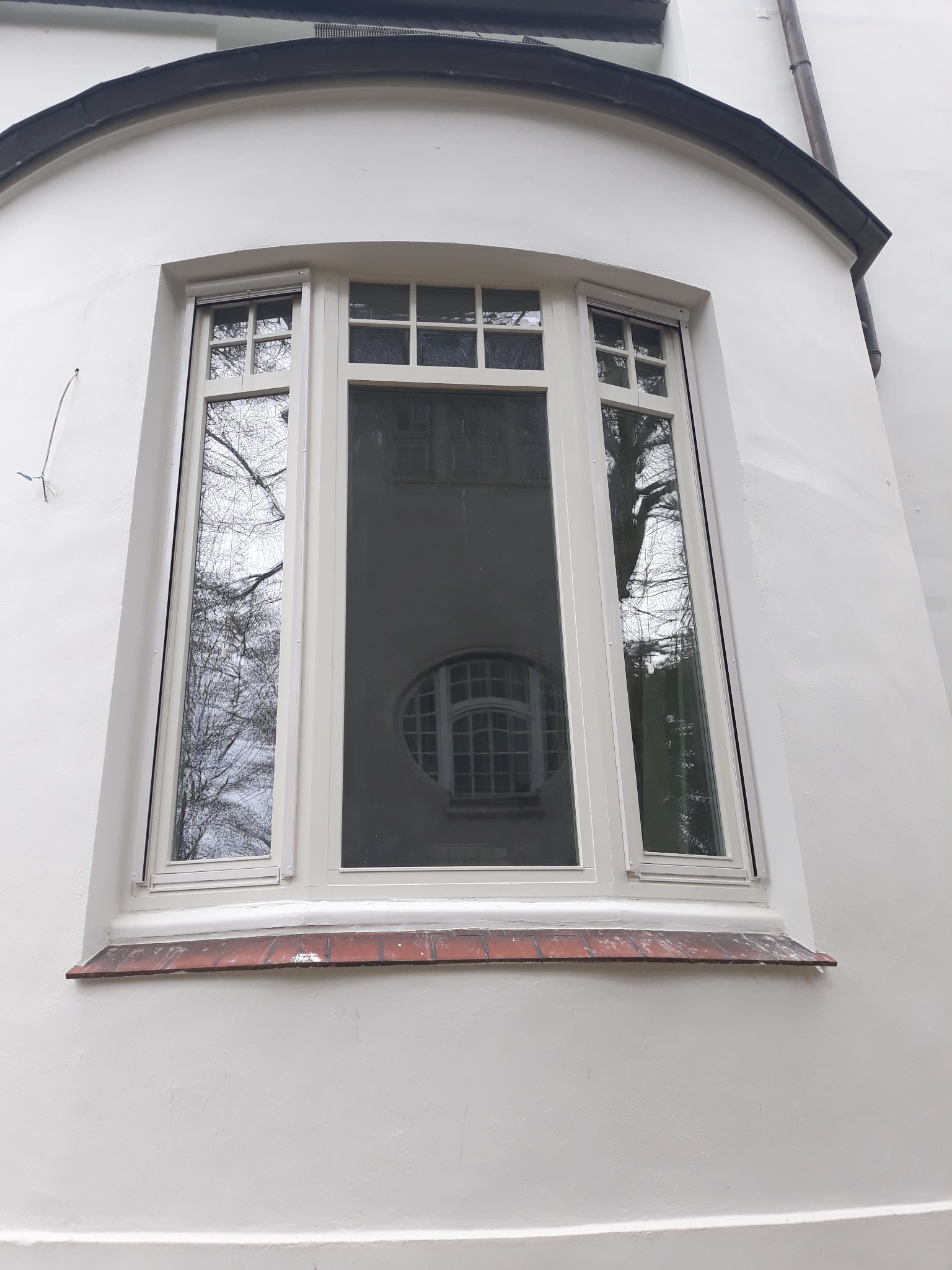 Białe okno z podziałami szprosowymi w budynku z białą elewacją, w szybach odbija się sąsiedni budynek z oknem w kształcie elipsy