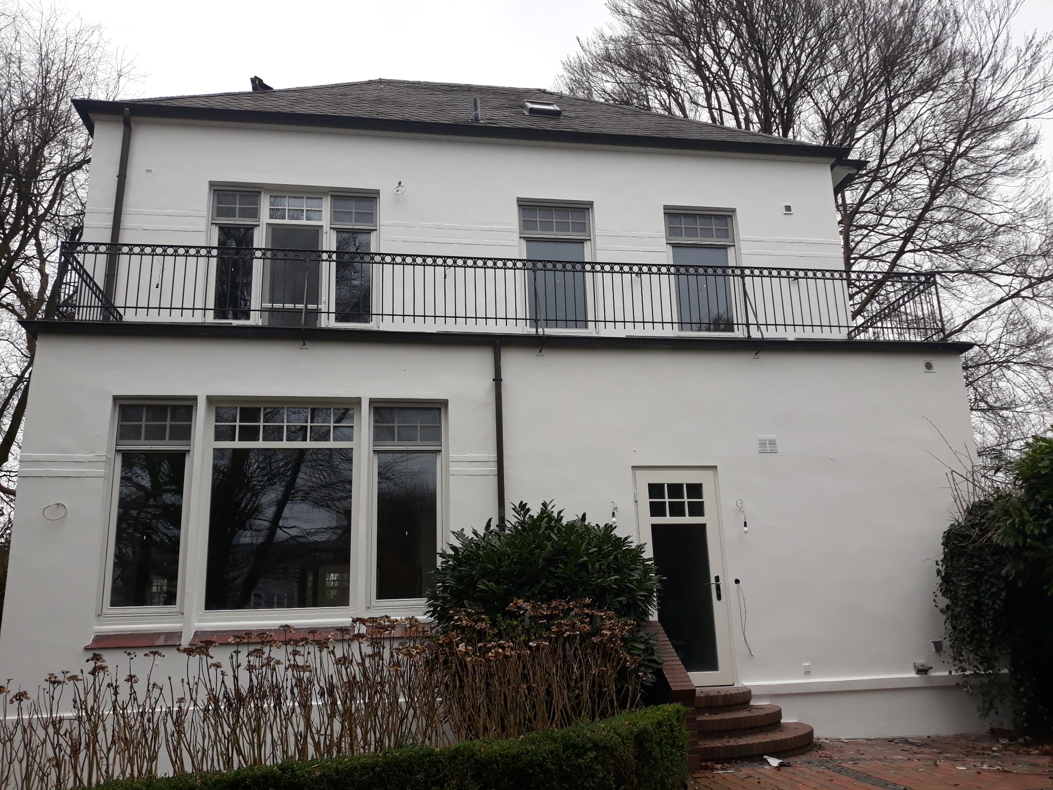 Biały, dwupiętrowy dom z balkonem i czarną balustradą, widoczne okna z białymi ramami, drzwi wejściowe z małymi szybkami i schodami, otoczony zielenią.