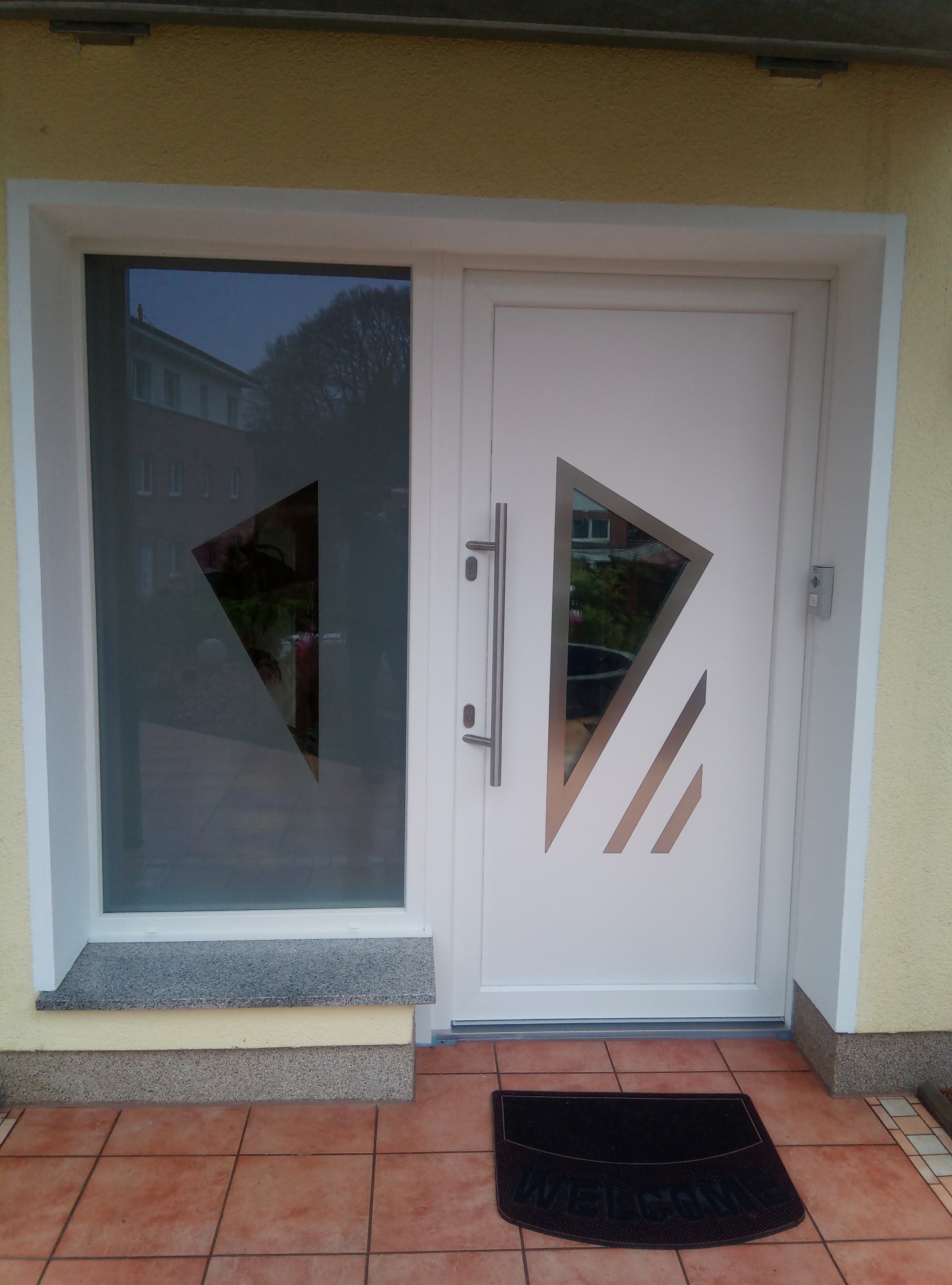 Białe drzwi wejściowe z geometrycznym wzorem i przeszkleniem, widoczny budynek odbijający się w szybie, wycieraczka 'WELCOME' na brązowych płytkach.