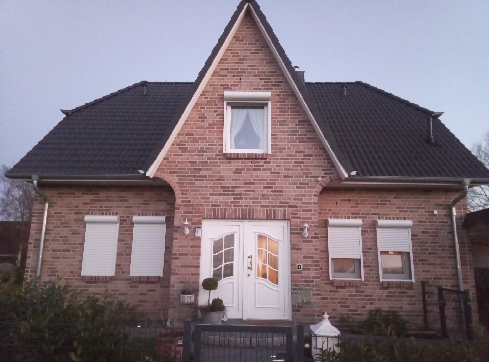 Dwukondygnacyjny dom z cegły z białymi drzwiami wejściowymi z szybami, częściowo opuszczonymi roletami na oknach i ciemną dachówką.