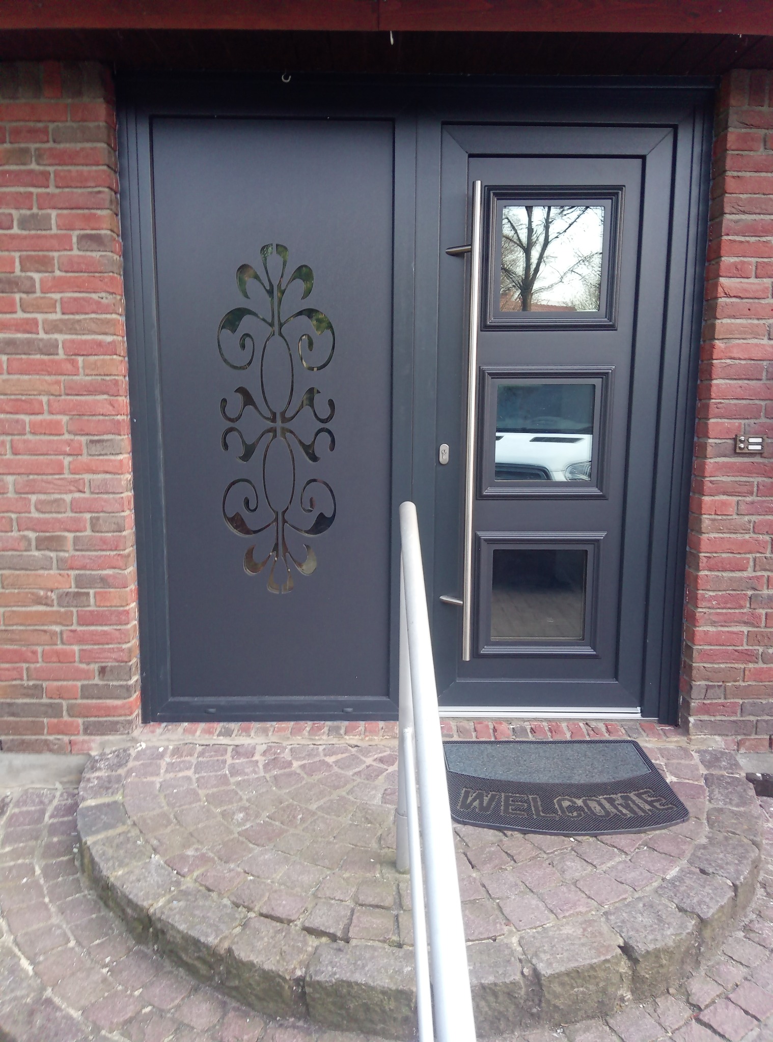 Grafitowe drzwi wejściowe z trzema małymi okienkami i ozdobnym wycięciem, widoczne z zewnątrz na tle ceglanej ściany i kamiennych schodów z wycieraczką.