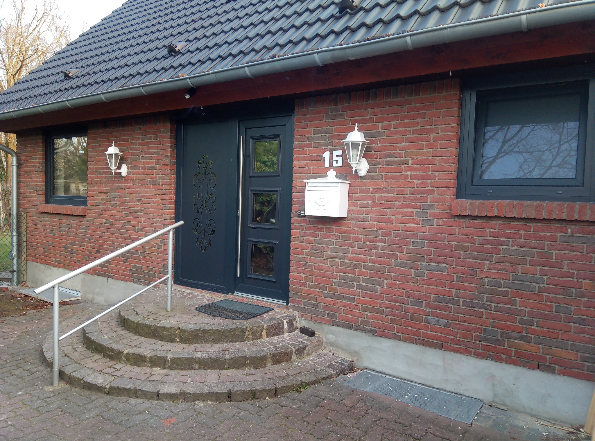 Dom z ciemnymi oknami PCV, drzwiami zewnętrznymi z dekoracyjnym wzorem, numerem domu '15' nad białą skrzynką pocztową i schodami z kostki brukowej z metalową poręczą.