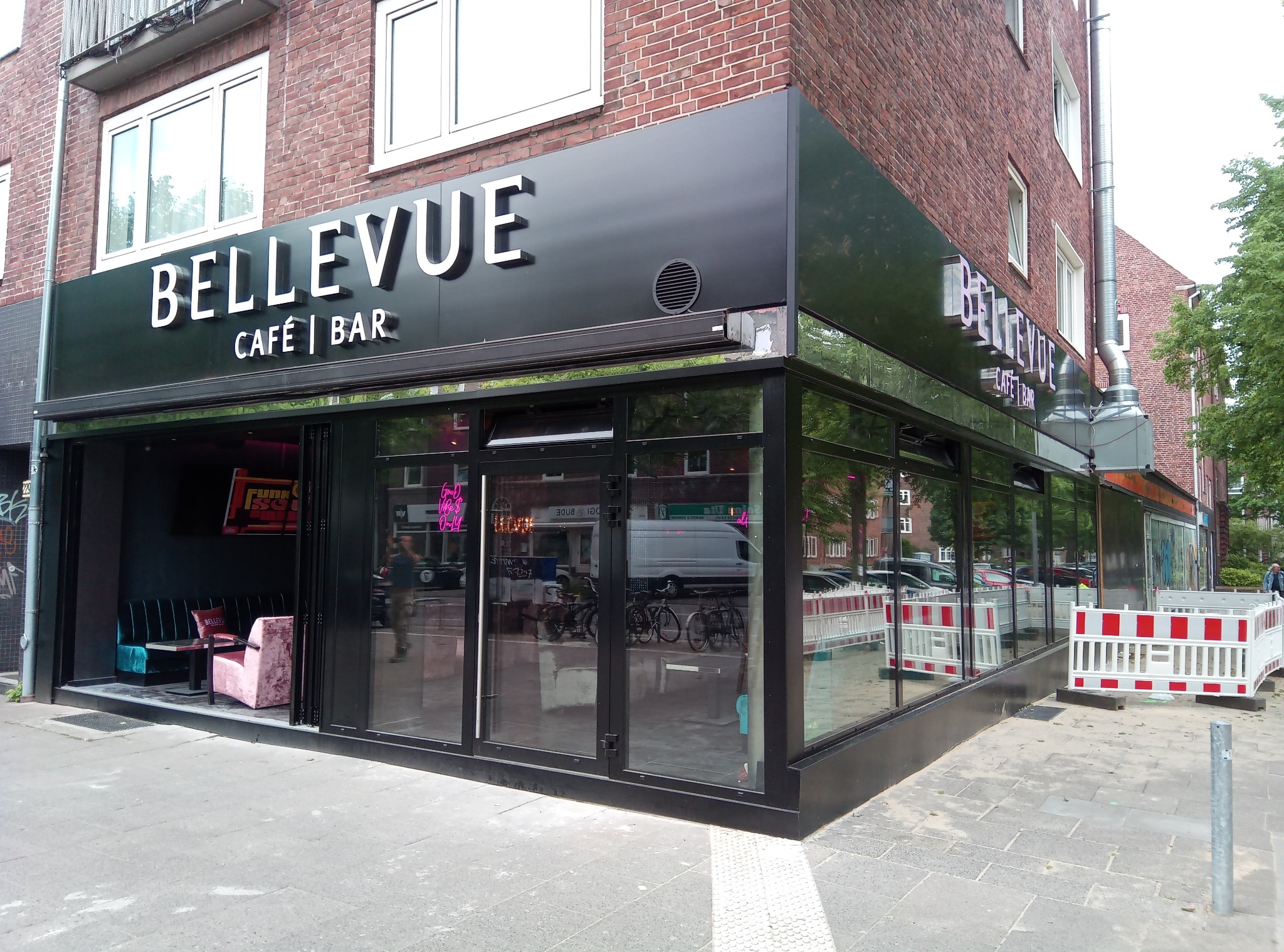 Fasada kawiarni Bellevue z dużymi, przeszklonymi przesuwnymi drzwiami w czarnej ramie, widoczne wnętrze z turkusową sofą i różowym fotelem, na zewnątrz barierki zabezpieczające.