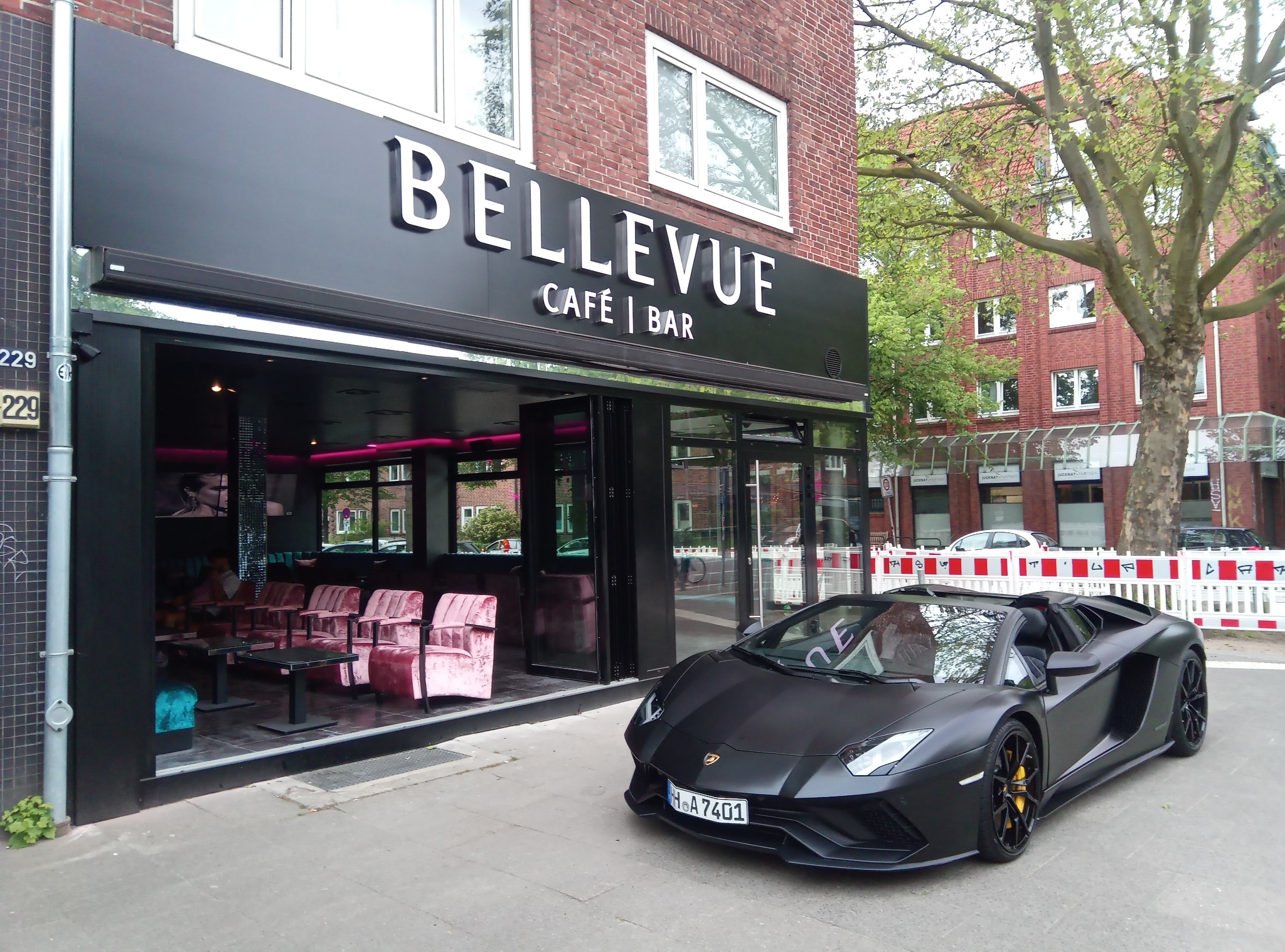 Eleganckie wejście do kawiarni Bellevue w Augustowie z czarną ramą i przeszklonymi drzwiami, obok zaparkowany matowy, czarny Lamborghini z żółtymi zaciskami hamulcowymi.