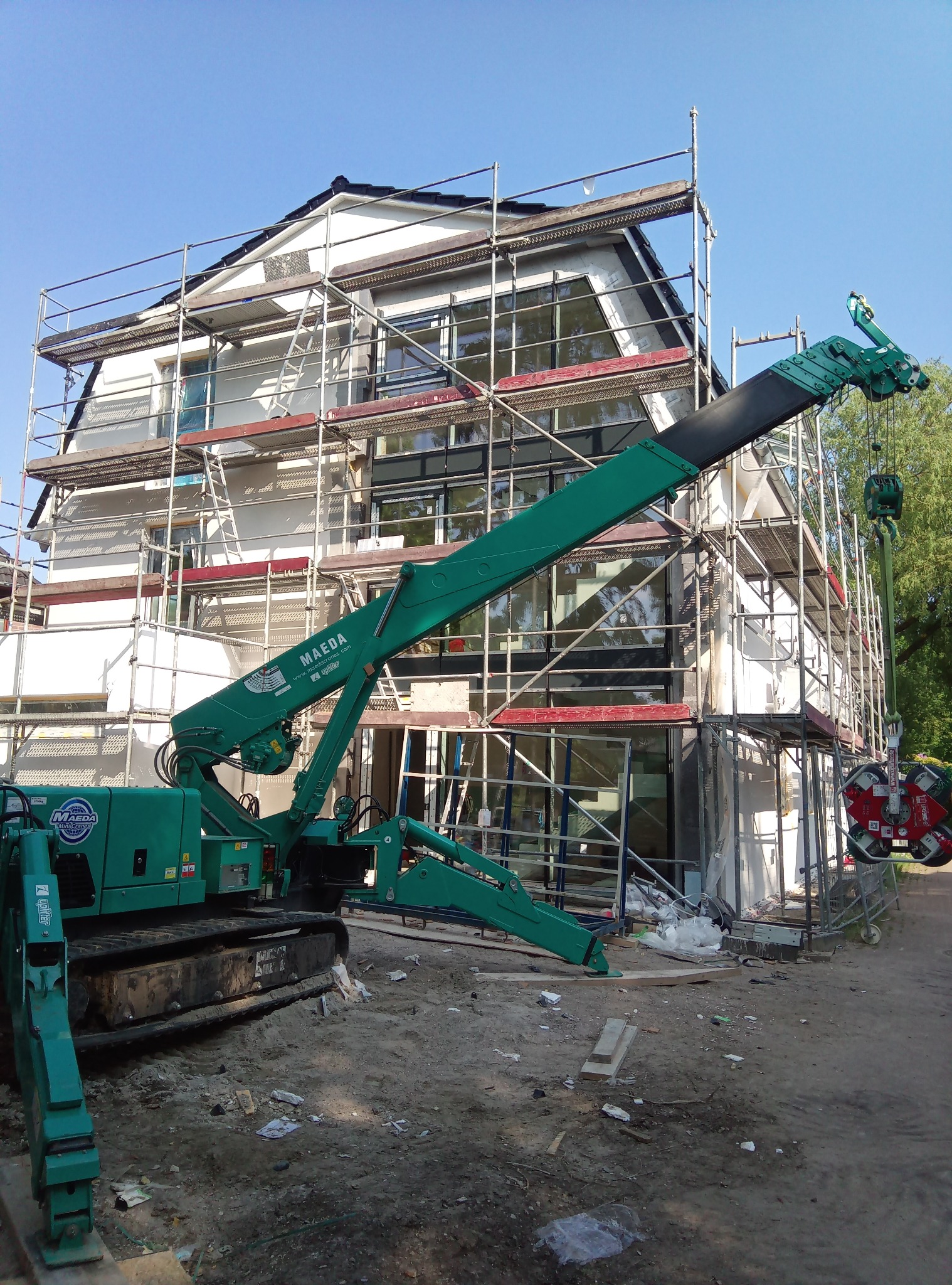 Montaż okien na rusztowaniu z użyciem zielonego dźwigu Maeda Mini Cranes, z przyssawkami do szkła, na tle dwupiętrowego domu w trakcie budowy.