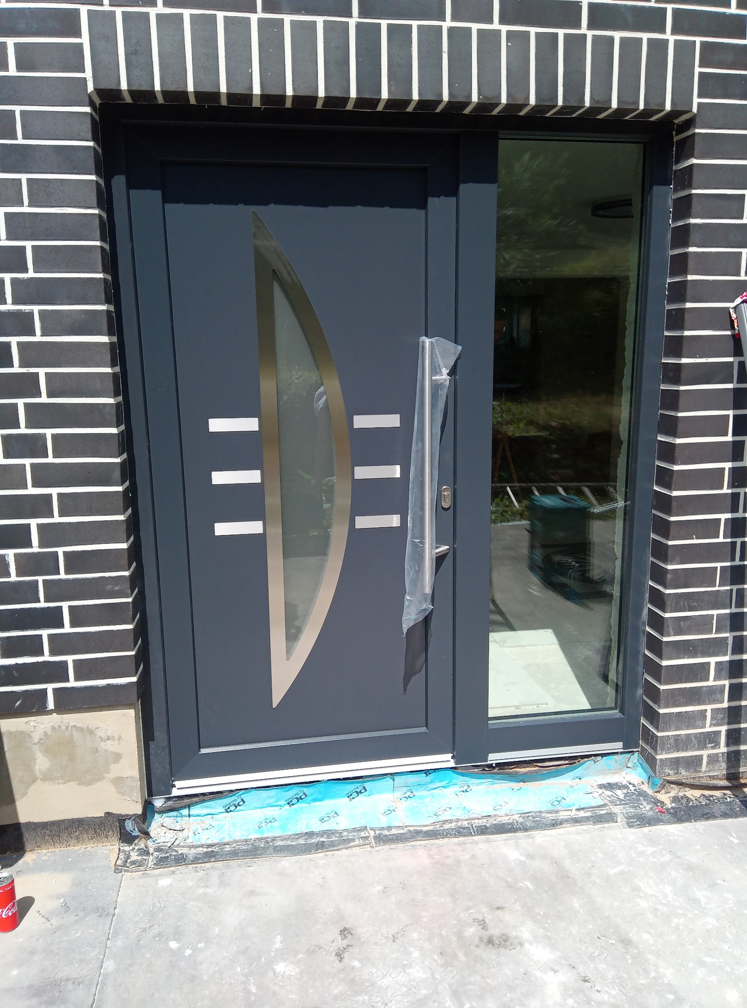 Szare drzwi wejściowe z szybą i ozdobnymi, poziomymi paskami, zamontowane w ścianie z czarnej cegły, uchwyt zabezpieczony folią, widoczny fragment podłoża z niebieską folią zabezpieczającą.