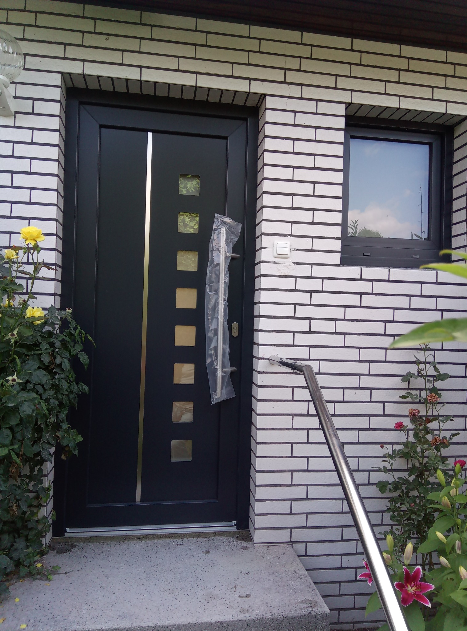 Nowoczesne, czarne drzwi wejściowe z kwadratowymi przeszkleniami i srebrnym akcentem, częściowo jeszcze w folii ochronnej, zamontowane w elewacji z białej cegły, widoczne schody wejściowe, kwiaty...
