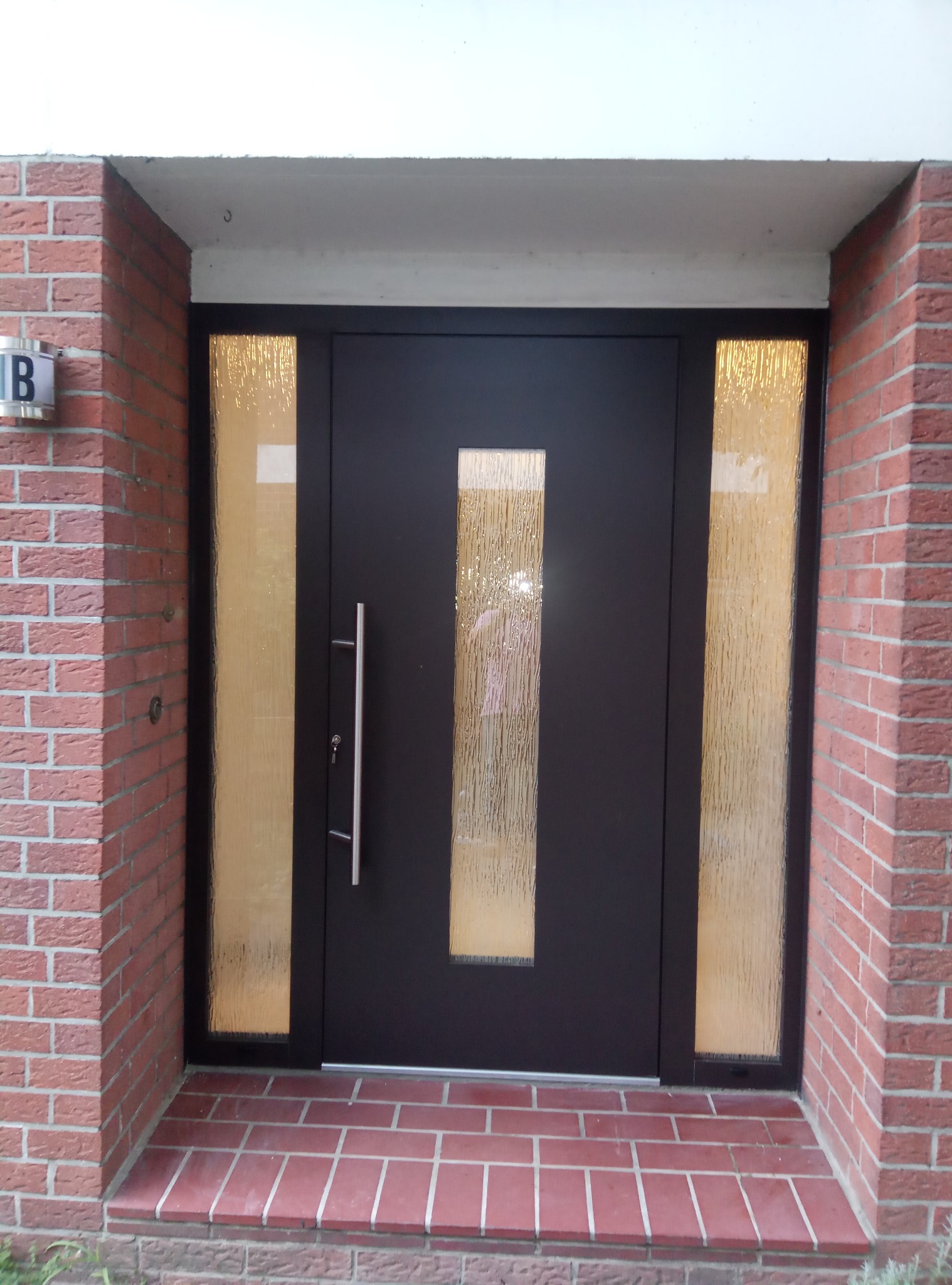 Ciemna, nowoczesna brama wejściowa z długim, pionowym uchwytem i szybami z ornamentowego szkła, wkomponowana w czerwoną ceglaną fasadę z numerem domu na boku.