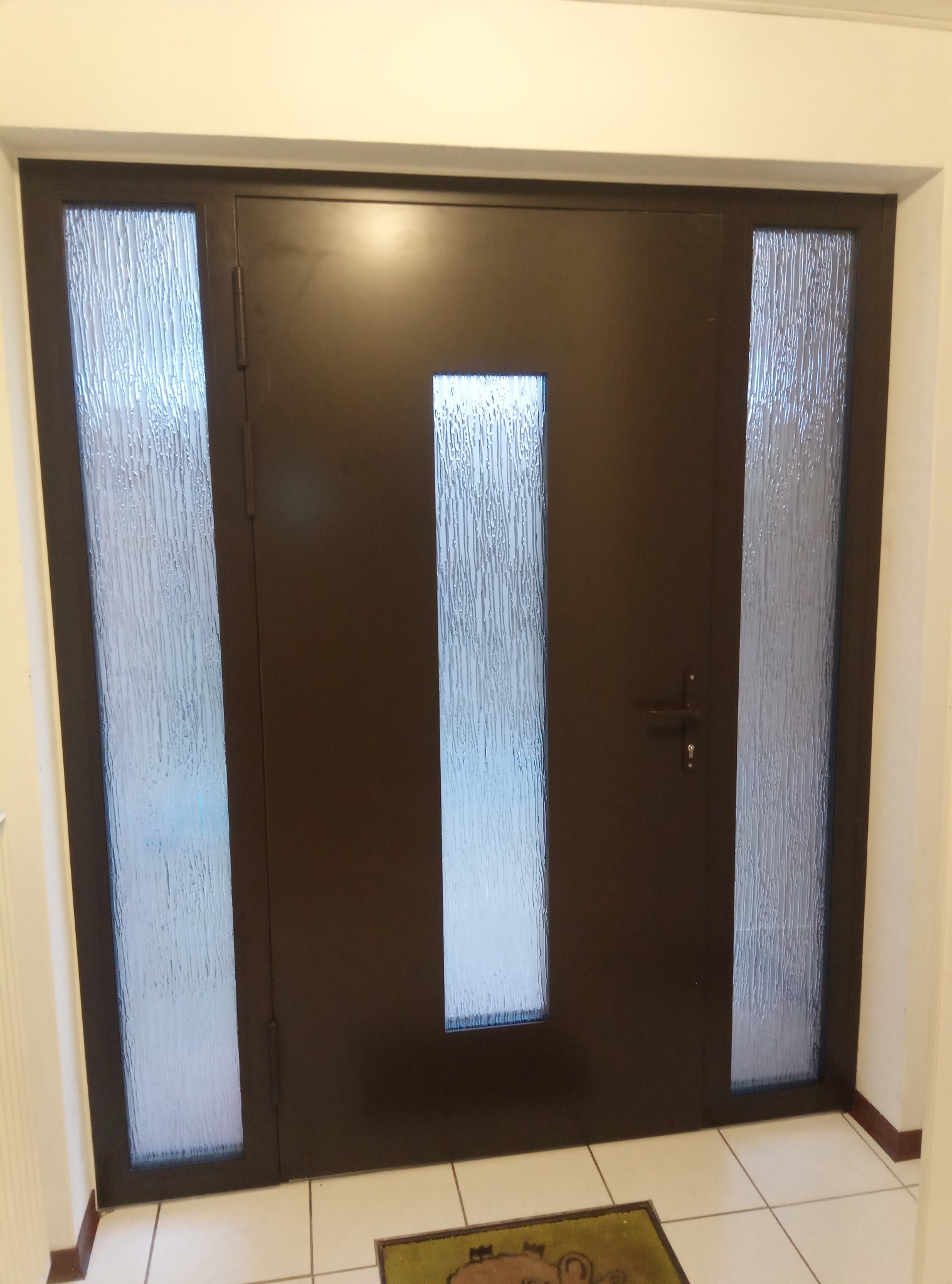 Ciemnobrązowe drzwi wejściowe z trzema szybami o fakturze kory drzewa, widoczne z bliska, z wycieraczką na kafelkowej podłodze.