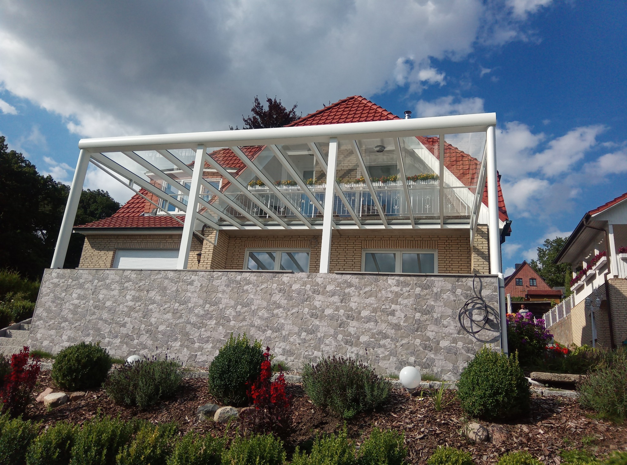 Zadaszenie tarasu z białym stelażem i przeszklonym dachem, widok z dołu na tle domu z czerwonym dachem i błękitnego nieba z chmurami.