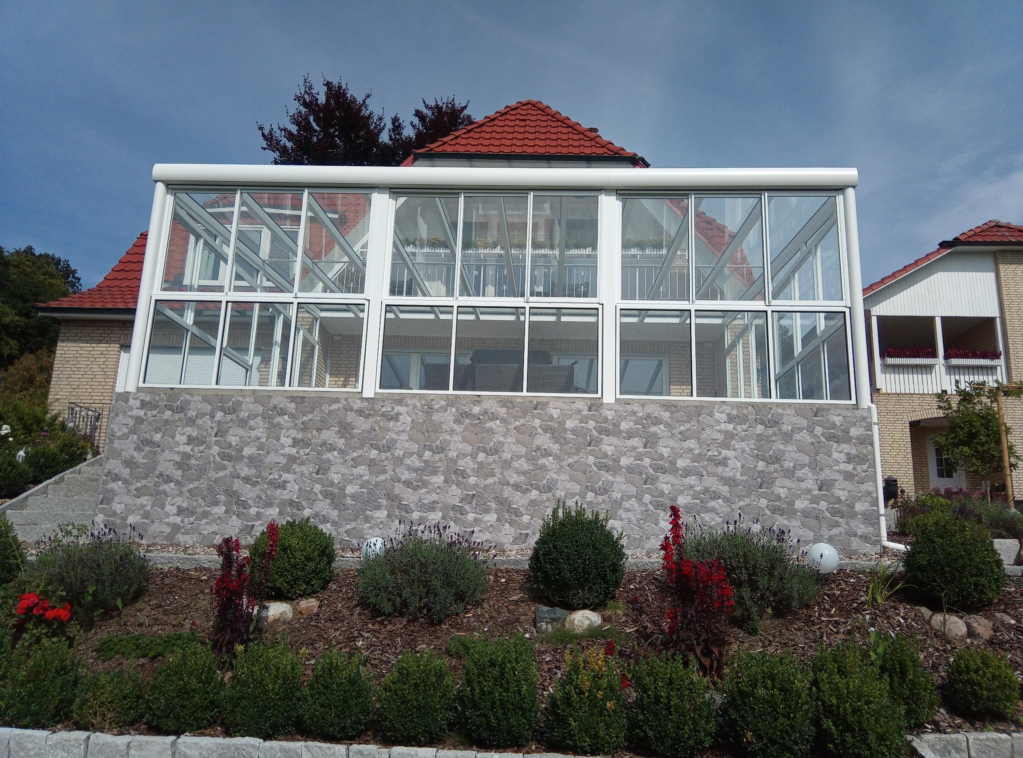Szklana zabudowa tarasu z białymi ramami na tle ceglanego domu z czerwonym dachem, widok z ogrodu z ozdobnymi krzewami i korą.