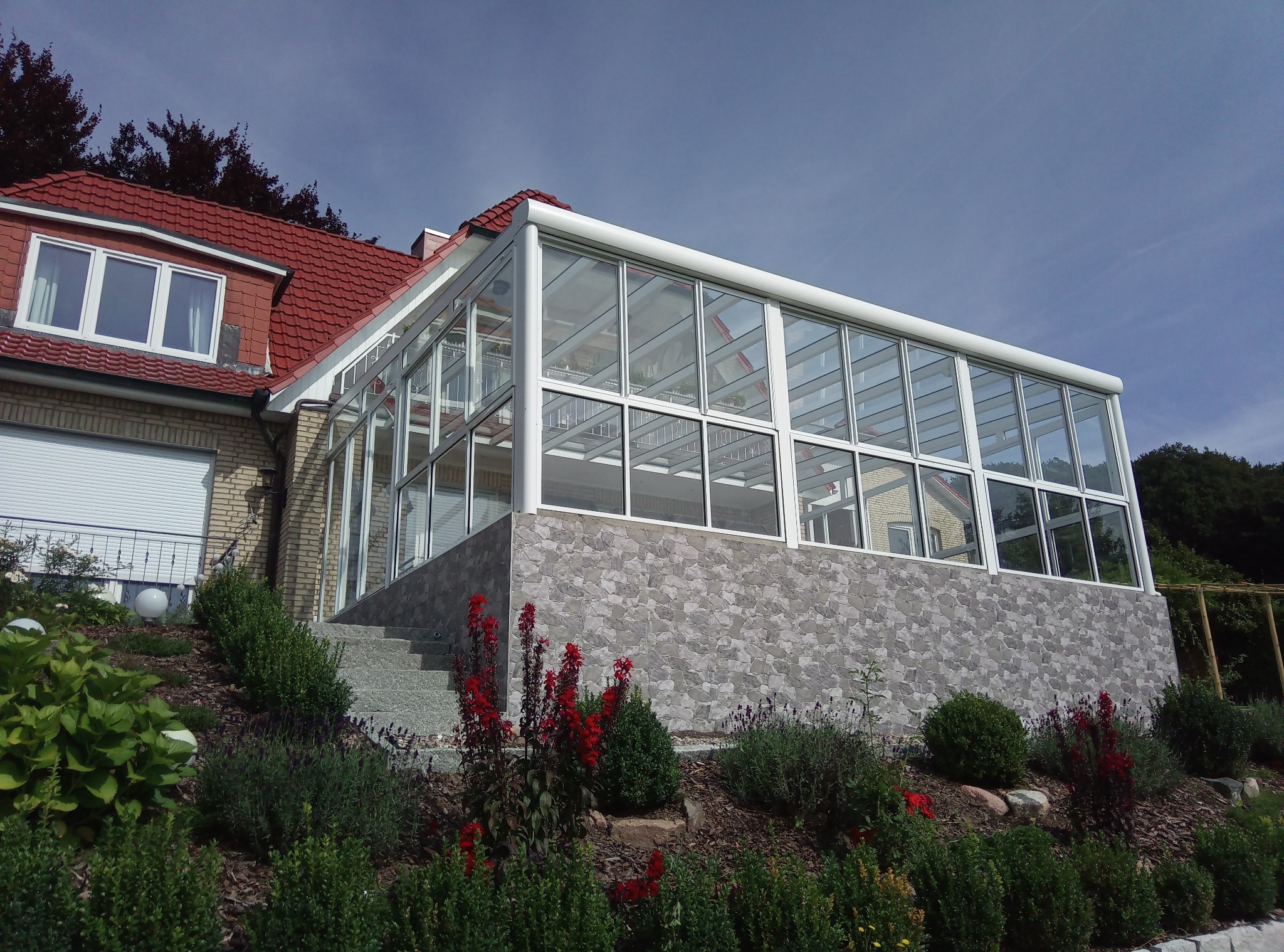 Szklana oranżeria przylegająca do domu z czerwonym dachem, widok z ogrodu z kwitnącymi czerwonymi i fioletowymi kwiatami na pierwszym planie.