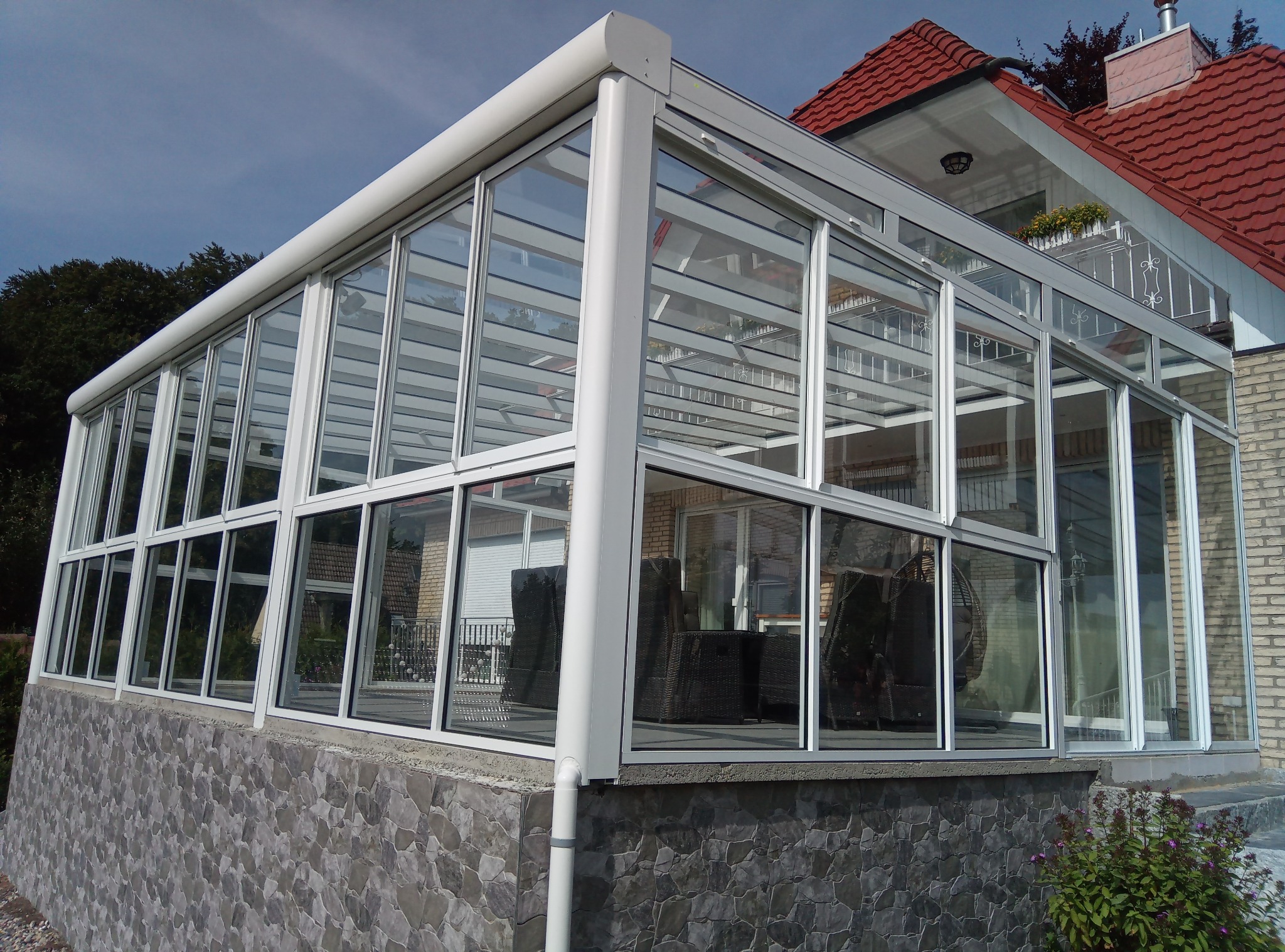 Zewnętrzny widok przeszklonej werandy z białymi ramami okiennymi, wewnątrz widoczne rattanowe meble ogrodowe, podmurówka wykończona kamieniem elewacyjnym.