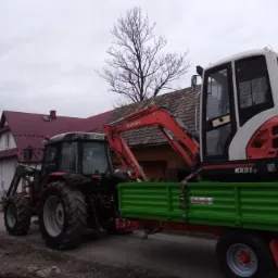 Czerwony traktor z ładowaczem transportuje na zielonej przyczepie biało-czerwoną mini koparkę Kubota KX91-3 na tle zabudowań wiejskich w pochmurny dzień.