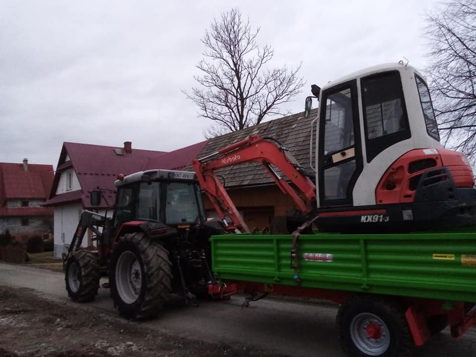Czerwony traktor z ładowaczem transportuje na zielonej przyczepie biało-czerwoną mini koparkę Kubota KX91-3 na tle zabudowań wiejskich w pochmurny dzień.