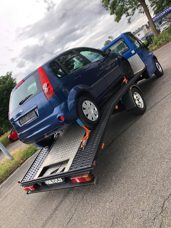 Niebieski Ford Fiesta zabezpieczony pomarańczowymi pasami na platformie lawety z metalową podłogą, widok z tyłu, tablica rejestracyjna widoczna.