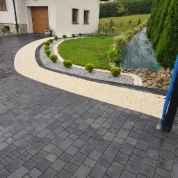 Elegancki podjazd z ciemnej kostki brukowej, obrzeża z jasnej kostki, ozdobne krzewy i trawnik przed nowoczesnym domem z garażem.