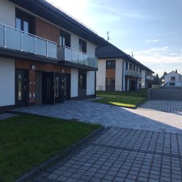 Firma Ogólnobudowlana OLEKSY Mariusz Oleksy - Nowoczesny budynek wielorodzinny z jasną elewacją, drewnianymi akcentami i szklanymi balkonami, widok z zewnątrz na kostkę brukową i trawnik.