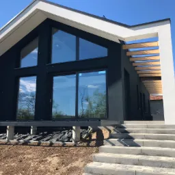 Nowoczesny dom z czarną elewacją, białymi akcentami i dużymi oknami, widoczne schody wejściowe i przygotowany teren wokół budynku.