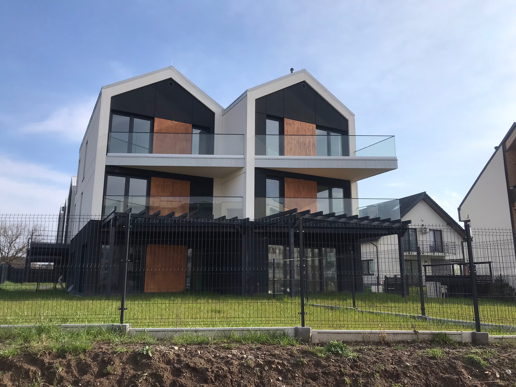 Nowoczesny dwupiętrowy bliźniak z balkonami o szklanych balustradach, wykończony białym tynkiem i panelami w kolorze drewna, otoczony czarnym metalowym ogrodzeniem.