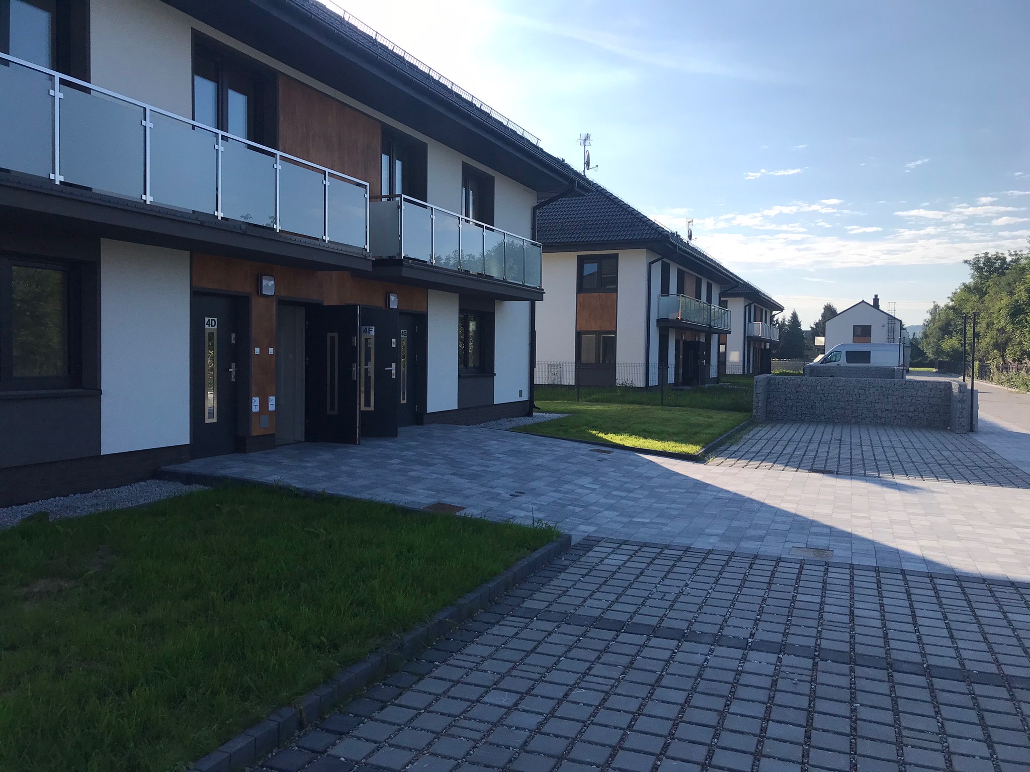 Nowoczesny budynek wielorodzinny z jasną elewacją, drewnianymi akcentami i szklanymi balkonami, widok z zewnątrz na kostkę brukową i trawnik.