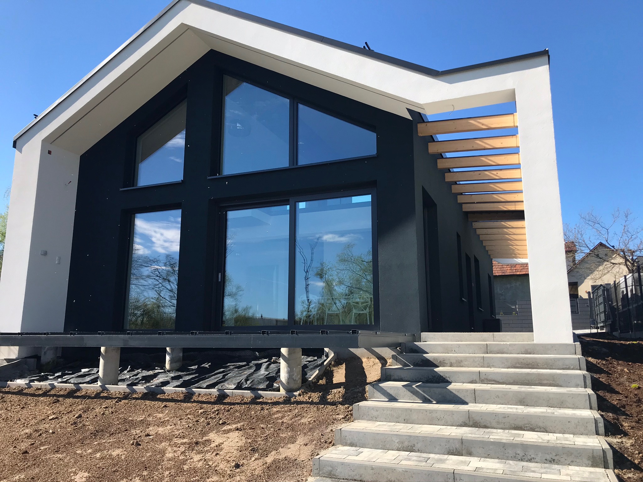 Nowoczesny dom z czarną elewacją, białymi akcentami i dużymi oknami, widoczne schody wejściowe i przygotowany teren wokół budynku.