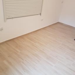 Usługi remontowo budowlane - Jasne, drewnopodobne płytki podłogowe ułożone w pustym pokoju z białymi ścianami, widoczne gniazdka elektryczne i okno z żaluzjami.
